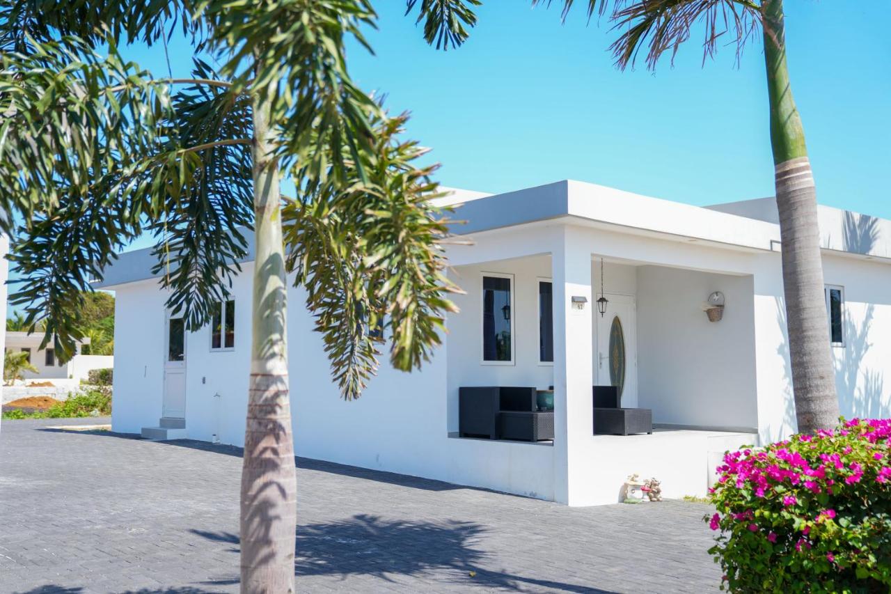 Modern white "Casa Gioia" villa with dark windows, covered porch, palm trees, and pink bougainvillea under a clear blue sky. Paved entrance.