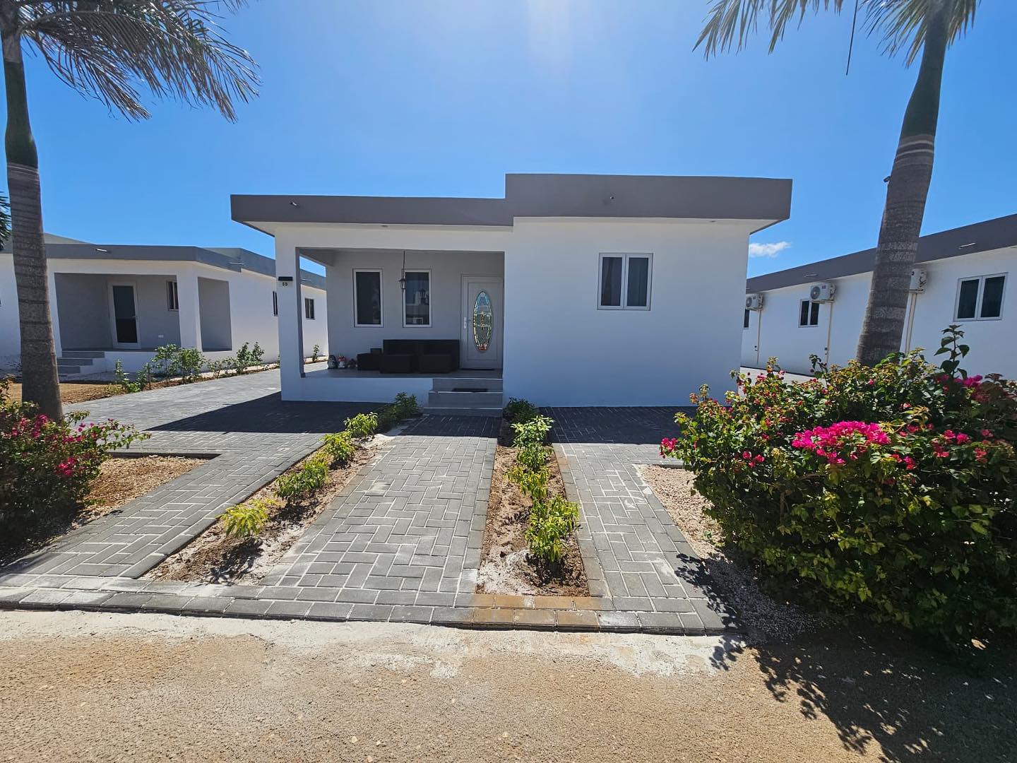Modern white "Casa Gioia" villa with dark windows, covered porch, palm trees, and pink bougainvillea under a clear blue sky. Paved entrance.